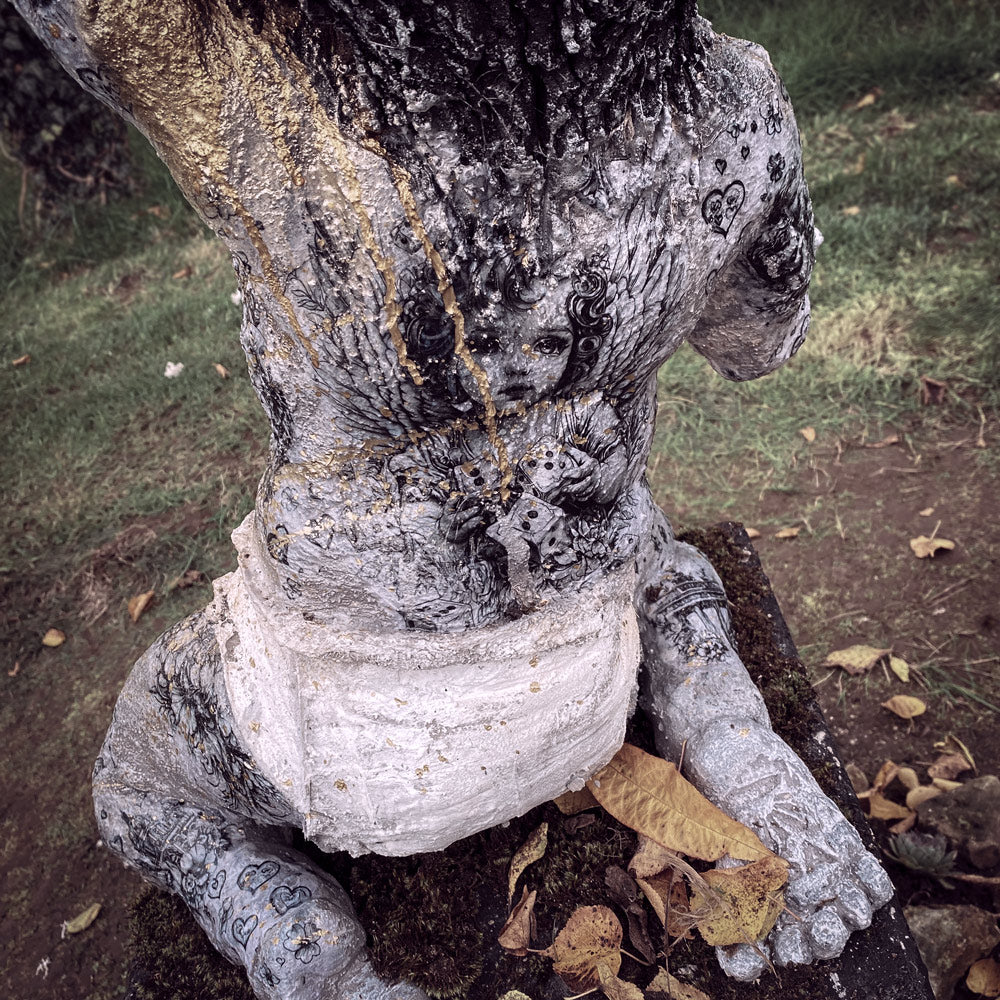 rückansicht von affenbaby statue aus beton mit einem großen engelchen bild aufseinem ganzen rücken - gold lack läuft über seine linke schulter herunter