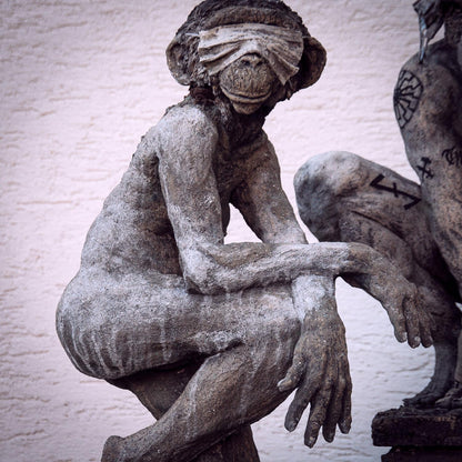 nackte affenfrauenskulptur mit einem fuss knienend, kann nix sehen weil sie mundschutz über den augen trägt und grinst