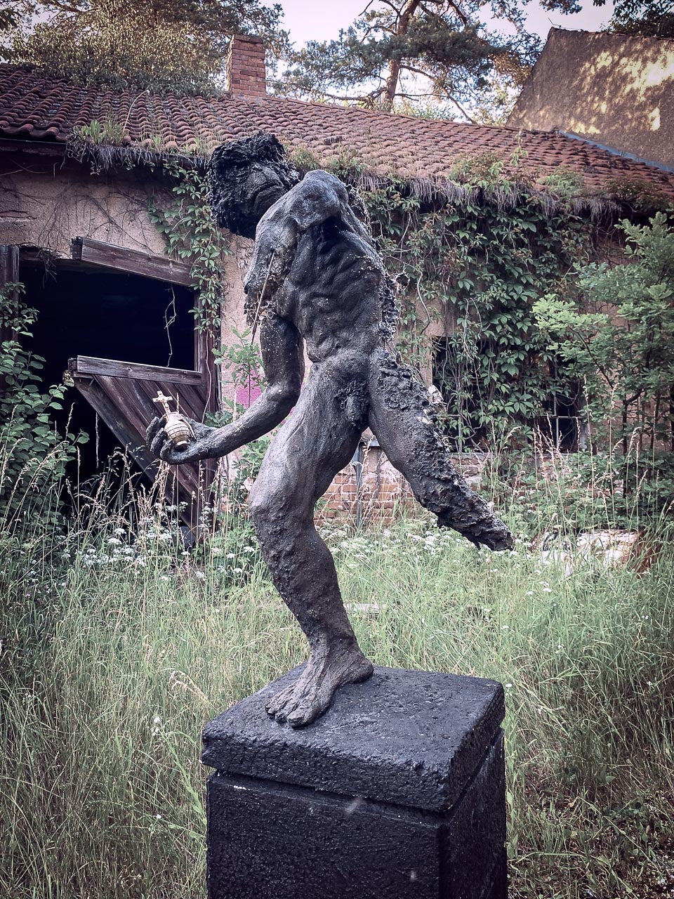 gesamtansicht von roland strallers iwan statue, der in  einer verlassenem von pflanzen überwucherten lostplace ruine, im hohen gras mit seiner heiligen goldenen handgranate aggressiv und halbzerszört posiert 