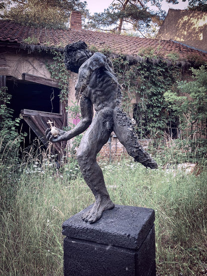 gesamtansicht von roland strallers iwan statue, der in  einer verlassenem von pflanzen überwucherten lostplace ruine, im hohen gras mit seiner heiligen goldenen handgranate aggressiv und halbzerszört posiert 