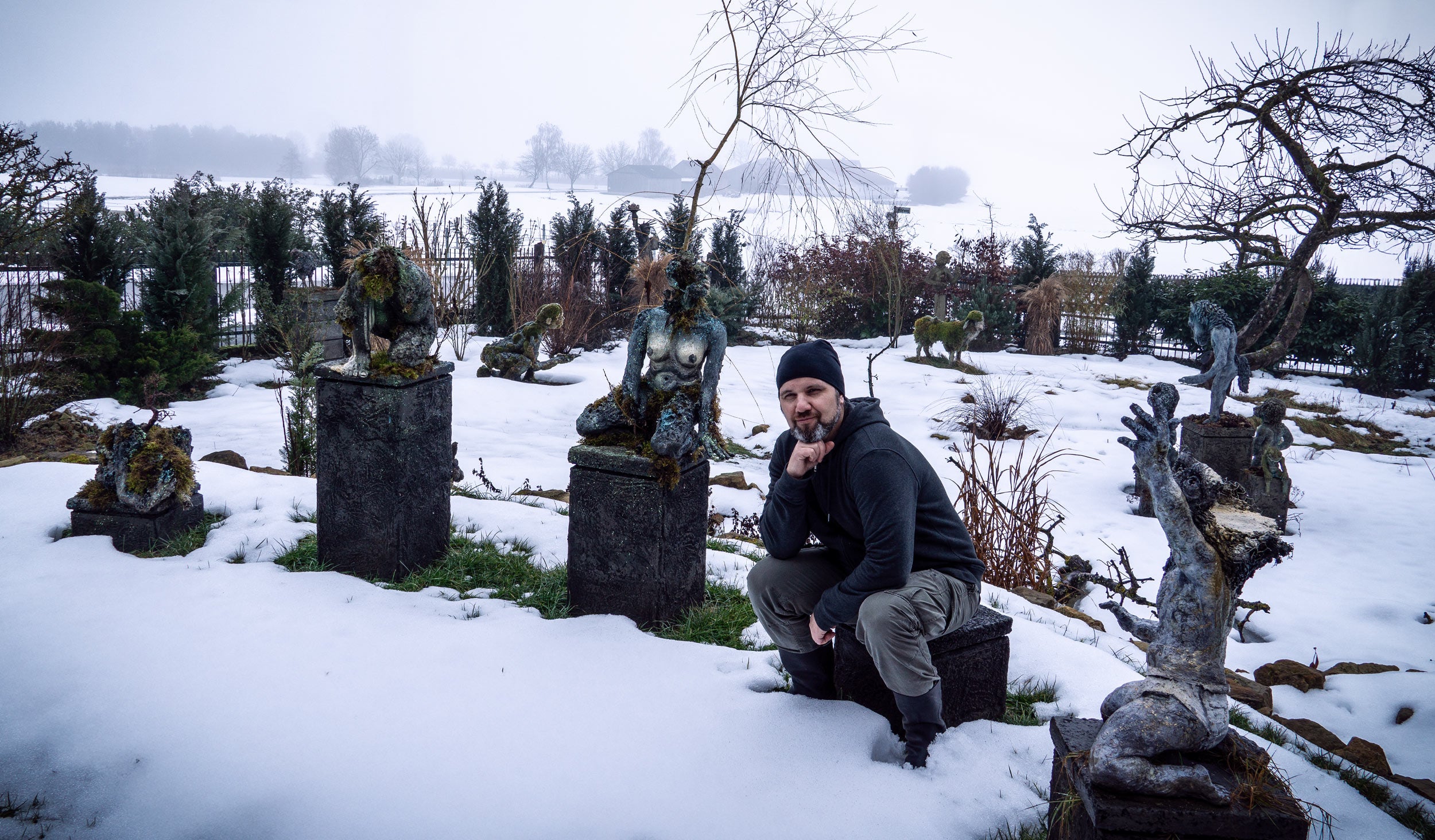 bildhauer roland straller sitzend in denkerpose zwischen seinen skulpturen im verschneiten garten - 
